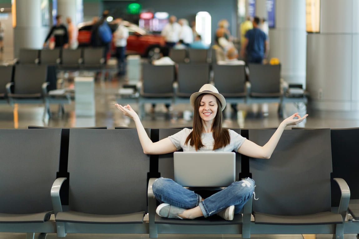 young-smiling-traveler-tourist-woman-with-laptop-sitting-with-crossed-legs-meditate-spread-hands-waiting-lobby-hall-airport.jpg
