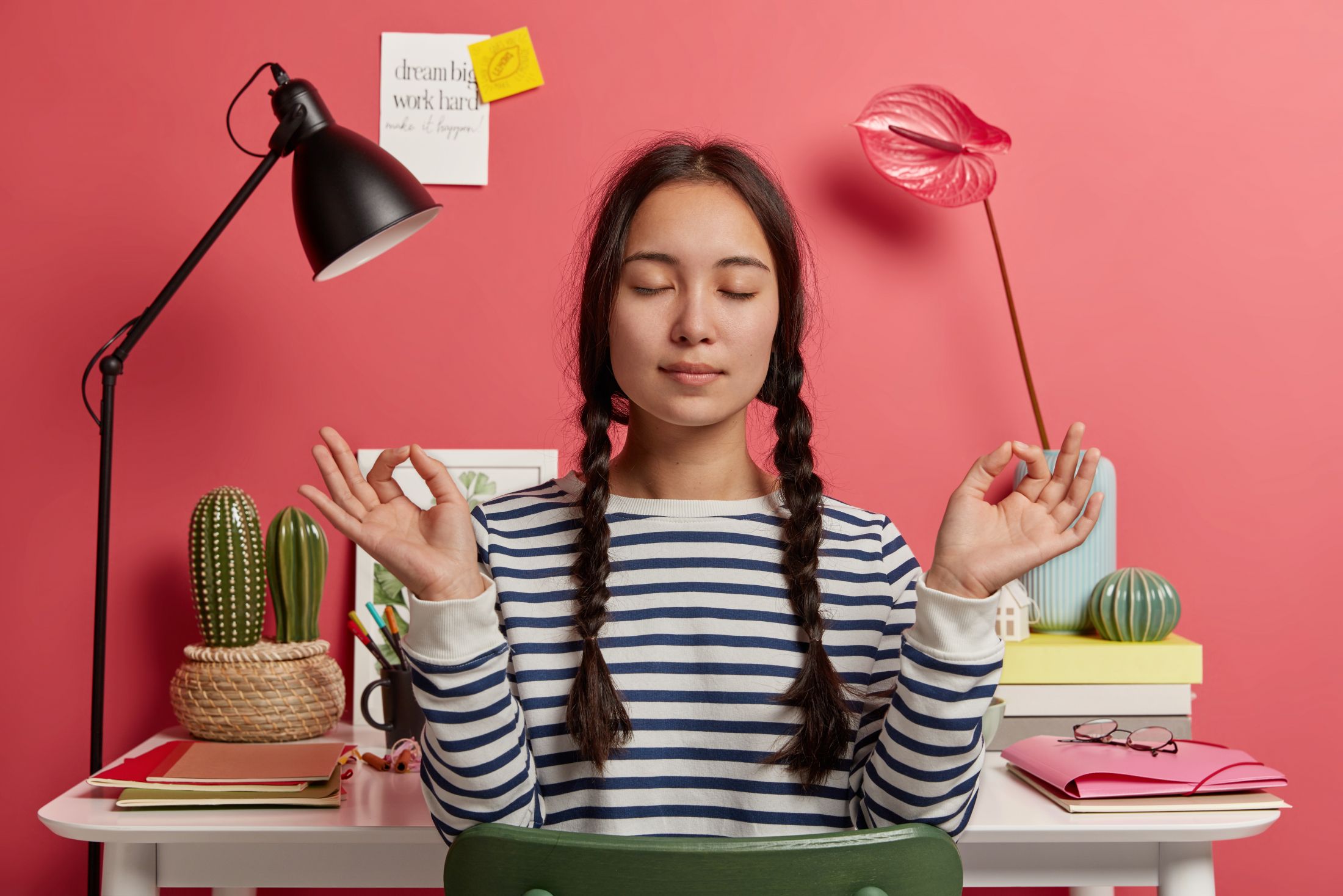 relaxed-asian-woman-meditates-at-workplace-sits-in-zen-pose-against-desktop-with-flowers-desk-lamp-notepads-wears-striped-casual-jumper-tries-to-relax-after-work-isolated-over-pink-background (1).jpg