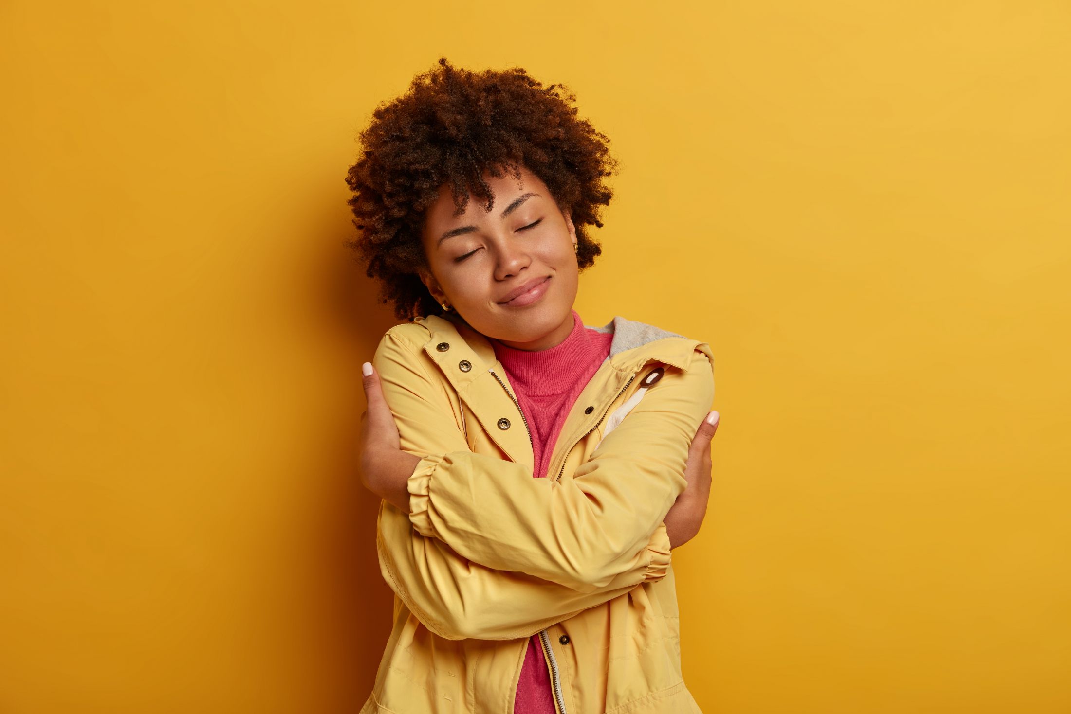selfishness-and-self-love-concept-portrait-of-pleased-dark-skinned-curly-female-model-hugs-herself-crosses-hands-over-body-keeps-eyes-closed-wears-jacket-poses-against-yellow-wall.jpg
